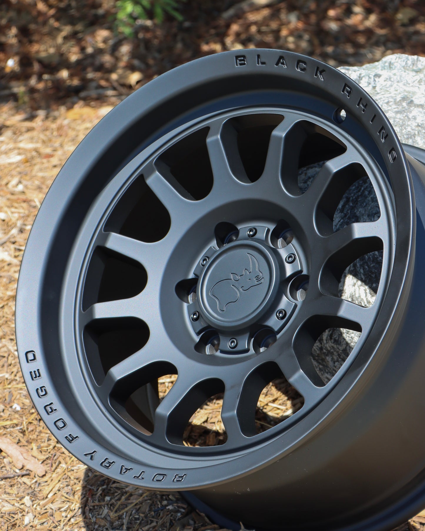 Black Rhino Rapid Wheel with a Matte Black finish, on the ground leaning against a rock.