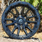 Fuel Rebel Wheel in a matte black finish, on the ground with a chain link fence in the background.