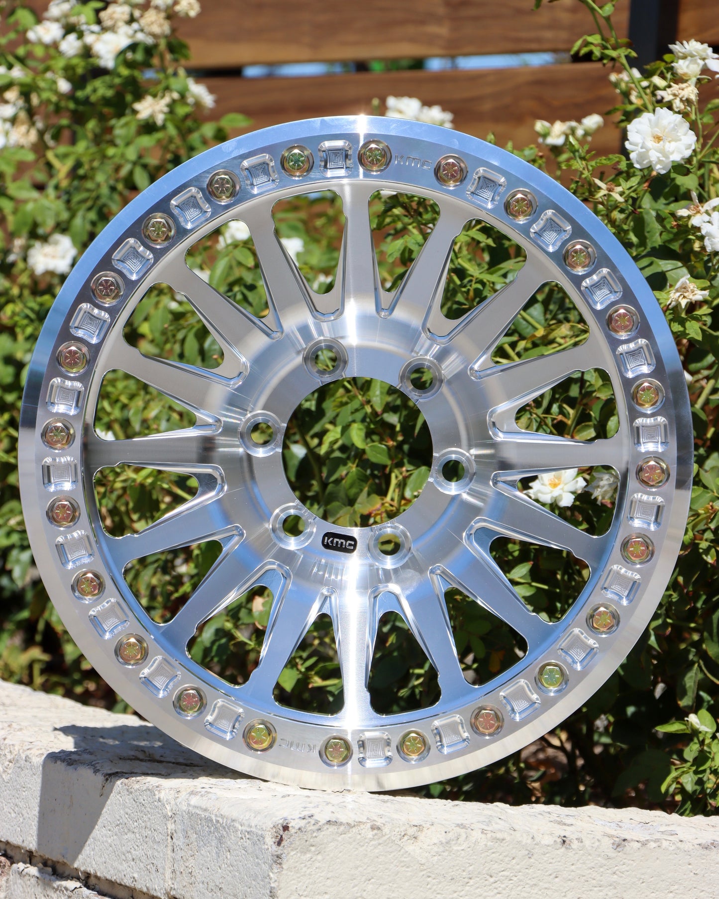 6 lug kmc impact forged bead lock wheel on the edge of a planter with bushes and a wood fence in the background.