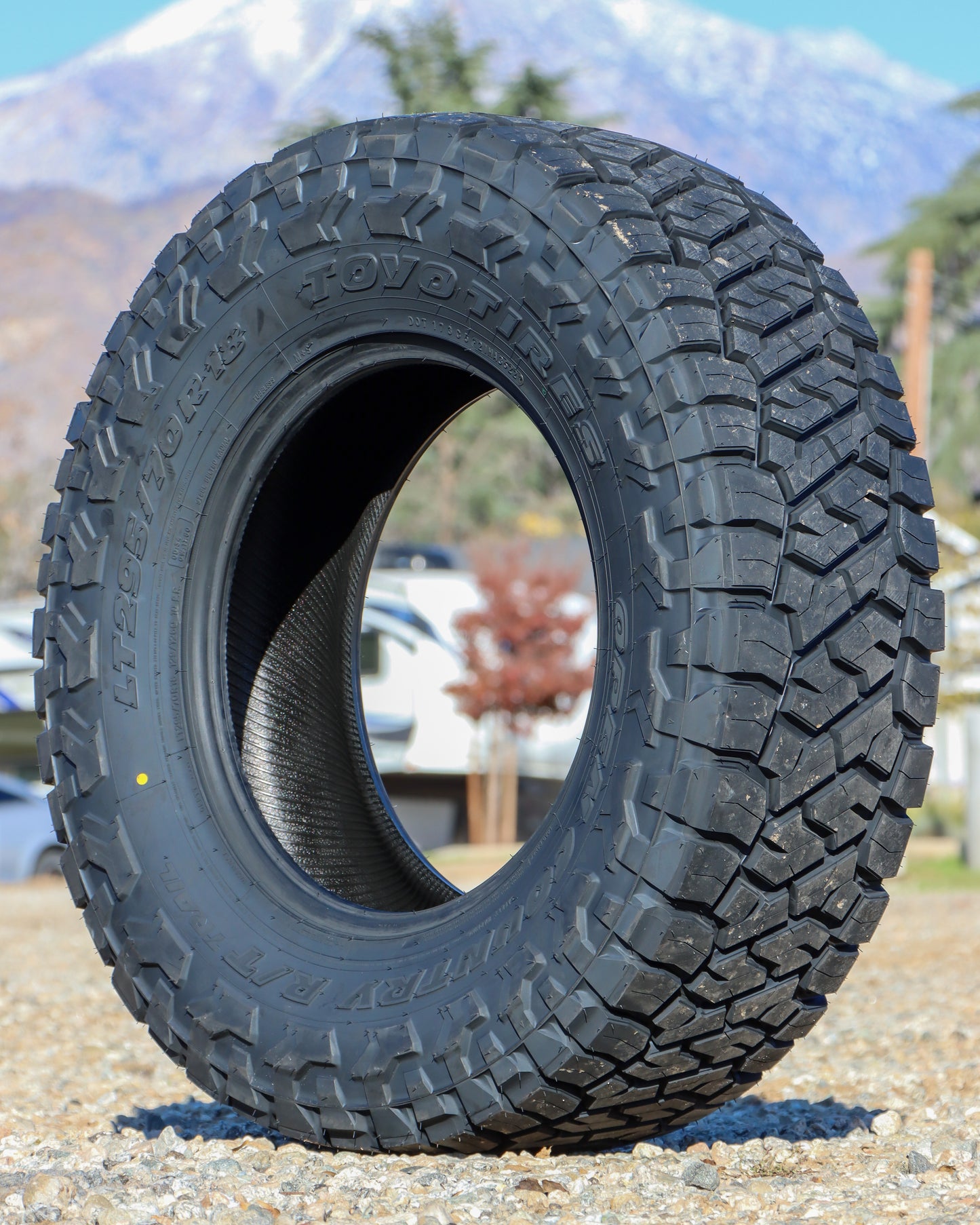 Toyo Open Country R/T Trail Tire sitting in a gravel field with trees and a snowy mountain in the background.