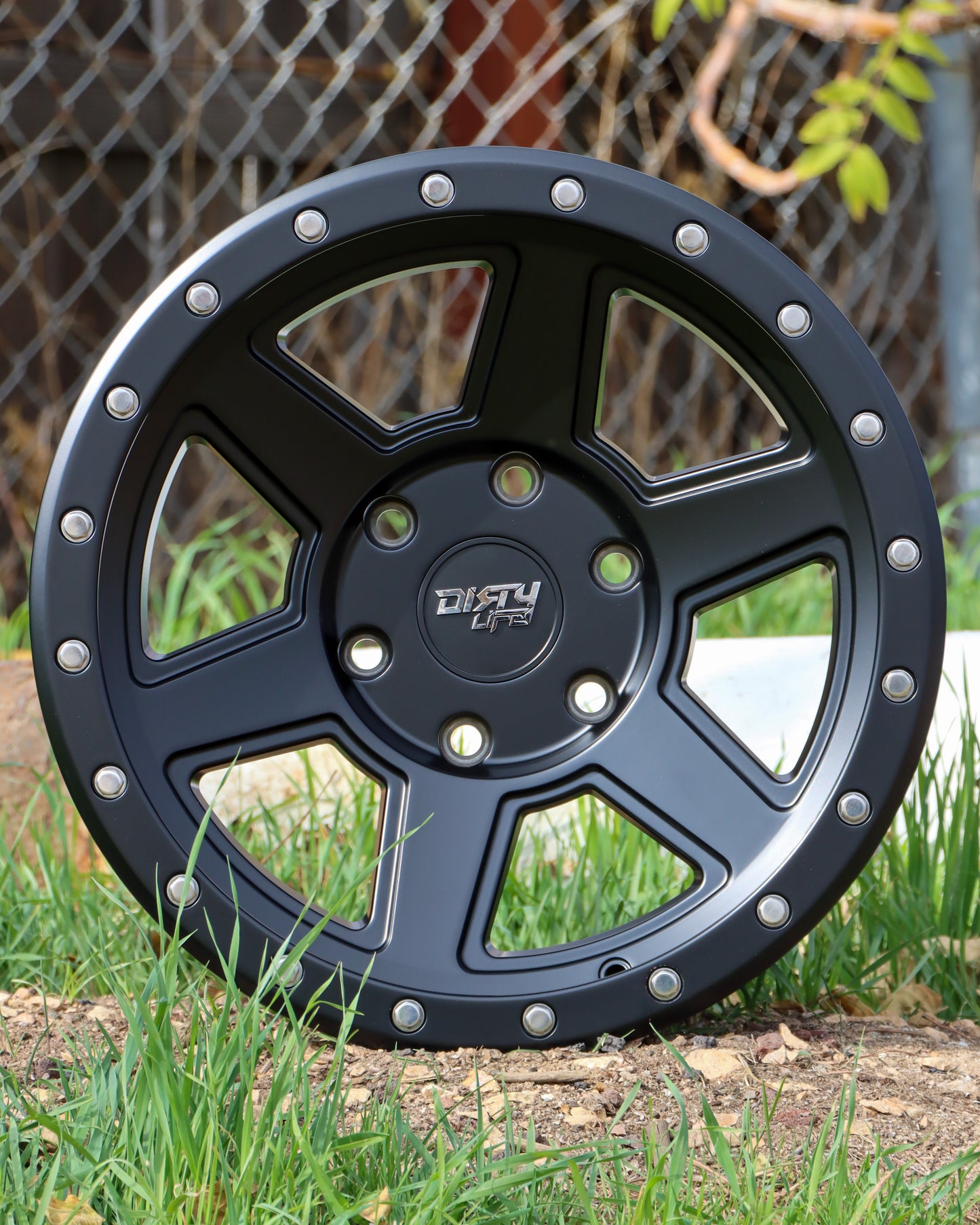 Dirty Life Compound Wheel Sitting in some dirt surrounded by grass, in a matte black finish.