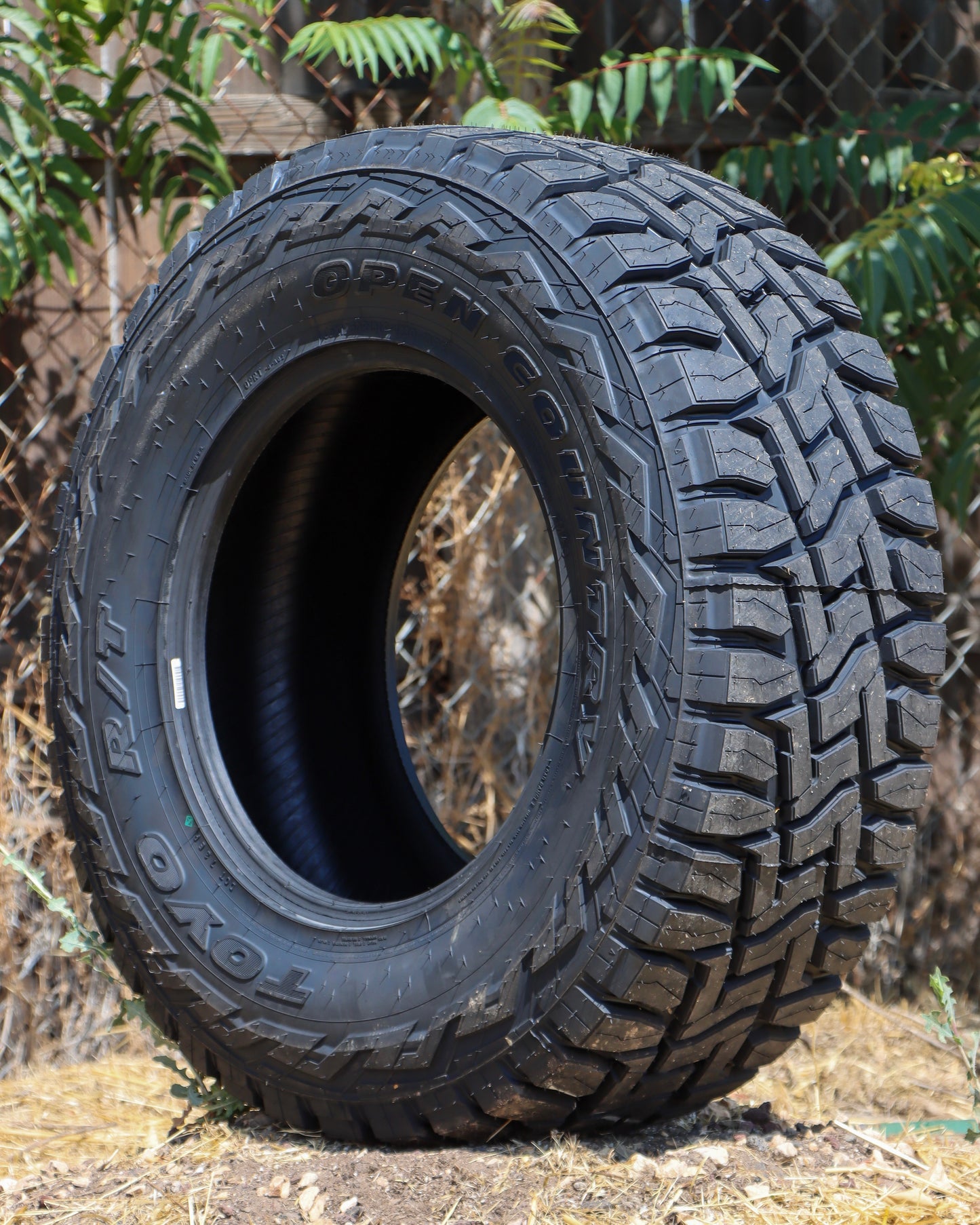 Toyo Open Country R/T tire sitting in a field with a chain link fence and trees in the background.