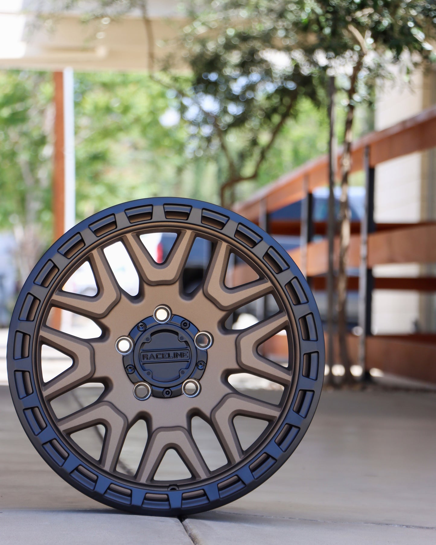Racline Krank Wheel in a Matte Bronze with a matte black ring, sitting on the concrete with a wood fence and little olive trees in the background.