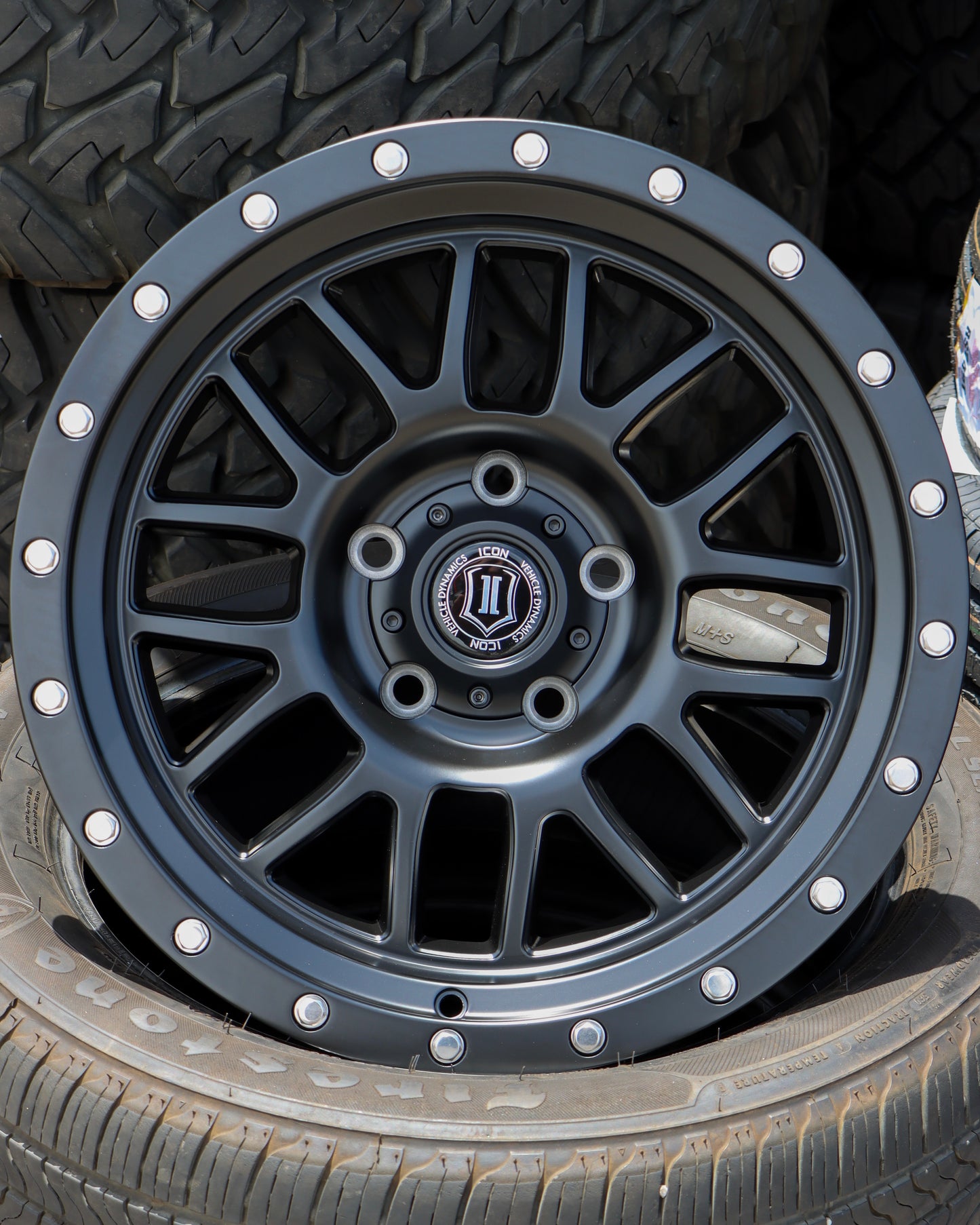 Icon Alpha wheel in a matte black finish, sitting in a tire with more tires in the backround.