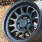 Black Rhino Rapid Wheel with a Matte Black finish, on the ground leaning against a rock.