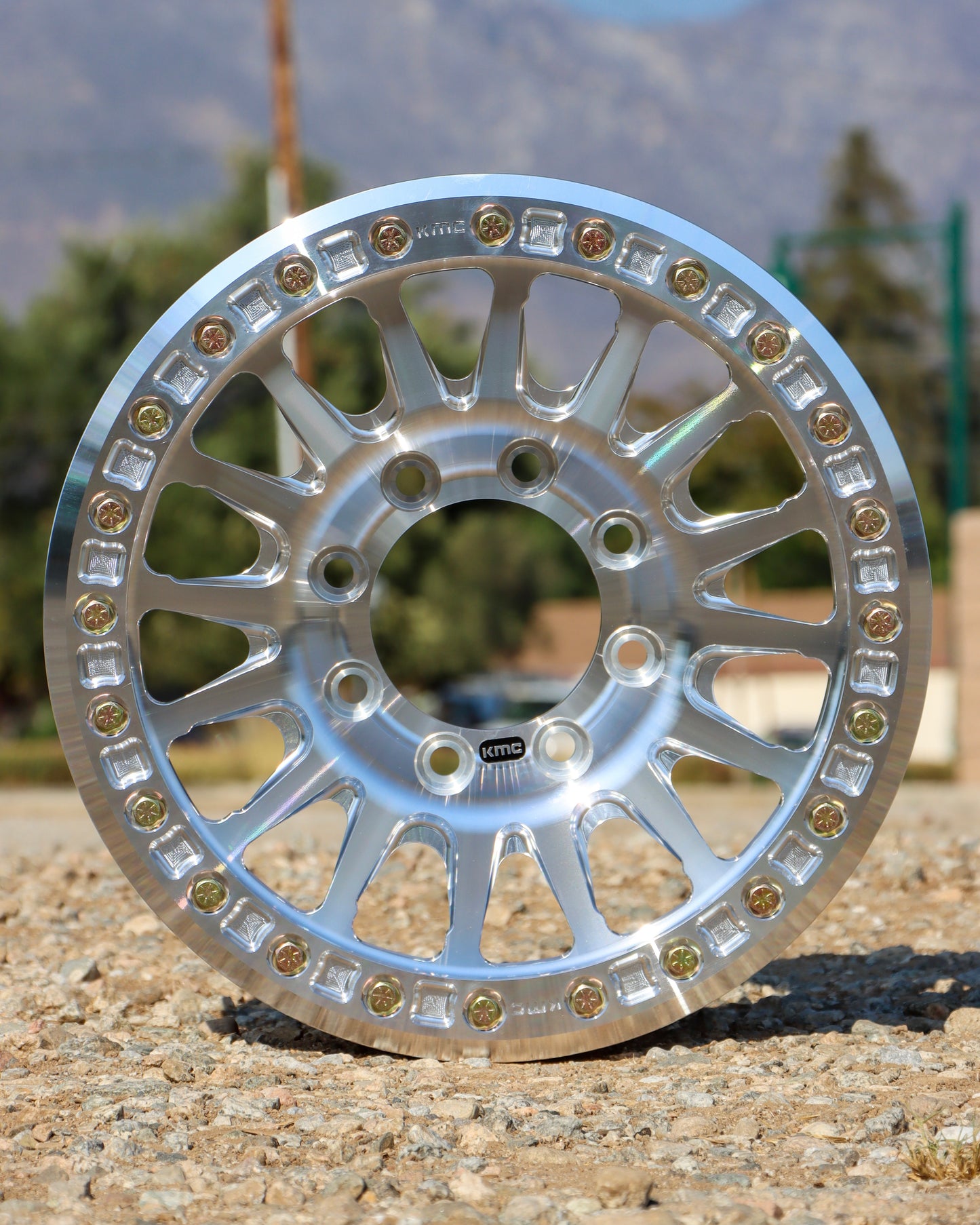 8 Lug KMC impact forged bead lock wheel sitting on some gravel with the mountains and trees in the background.