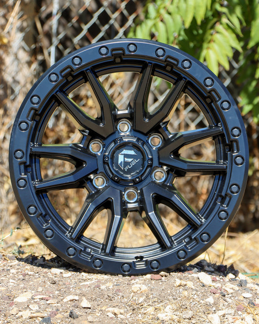 Fuel Rebel Wheel in a matte black finish, on the ground with a chain link fence in the background.
