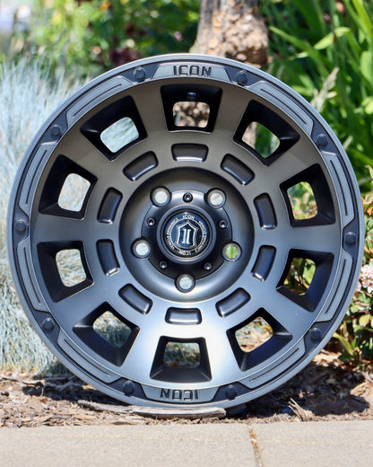 Icon Thrust Wheel in a smoked satin black finish sitting in a planter with a tree stump and bushes in the background.