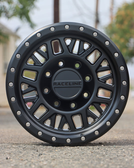 Raceline Ryno Wheel in a matte black finish sitting in the middle of an alley with trees in the background.