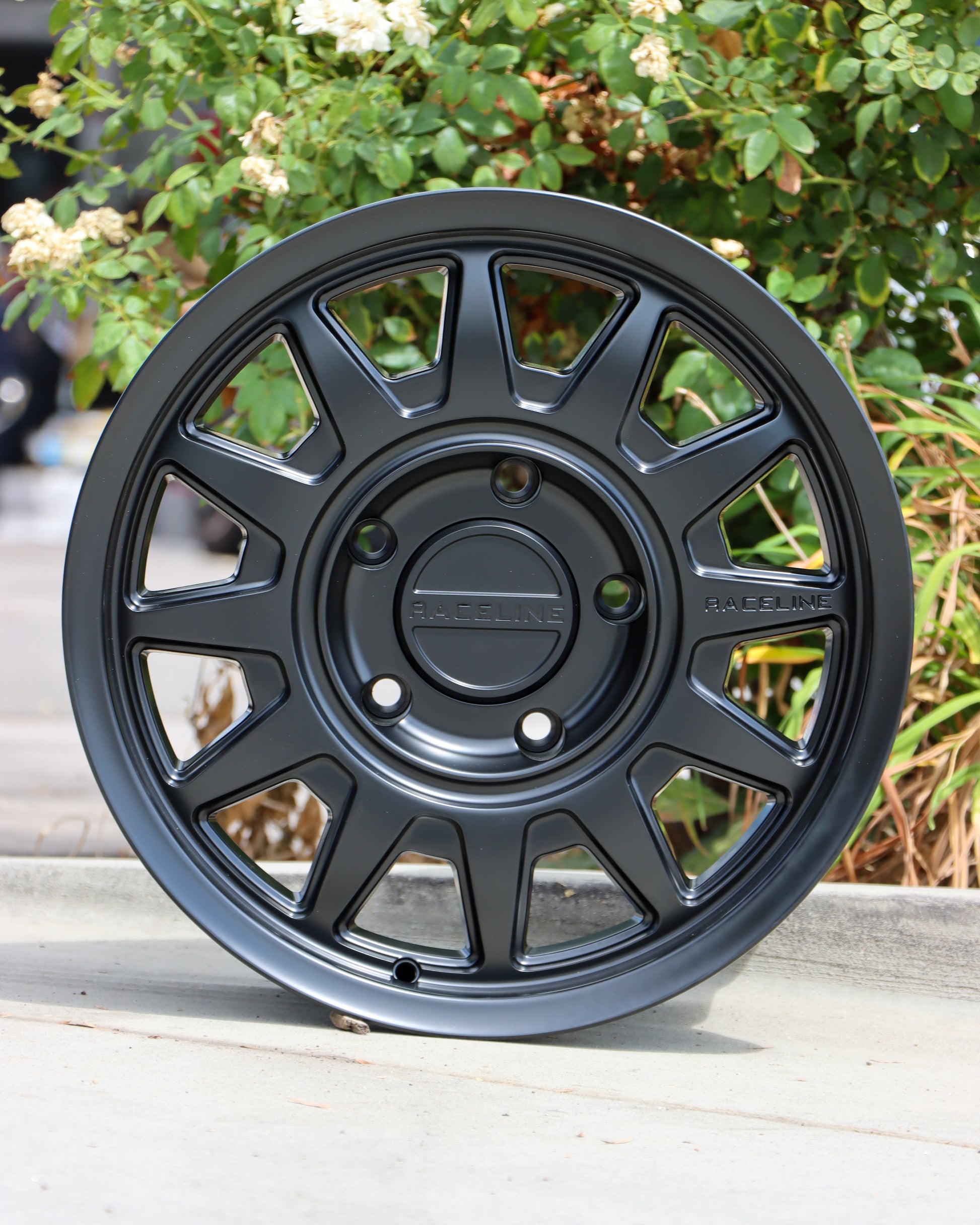 Raceline Areo wheel in a matte black finish sitting on the sidewalk with bushes in the background.