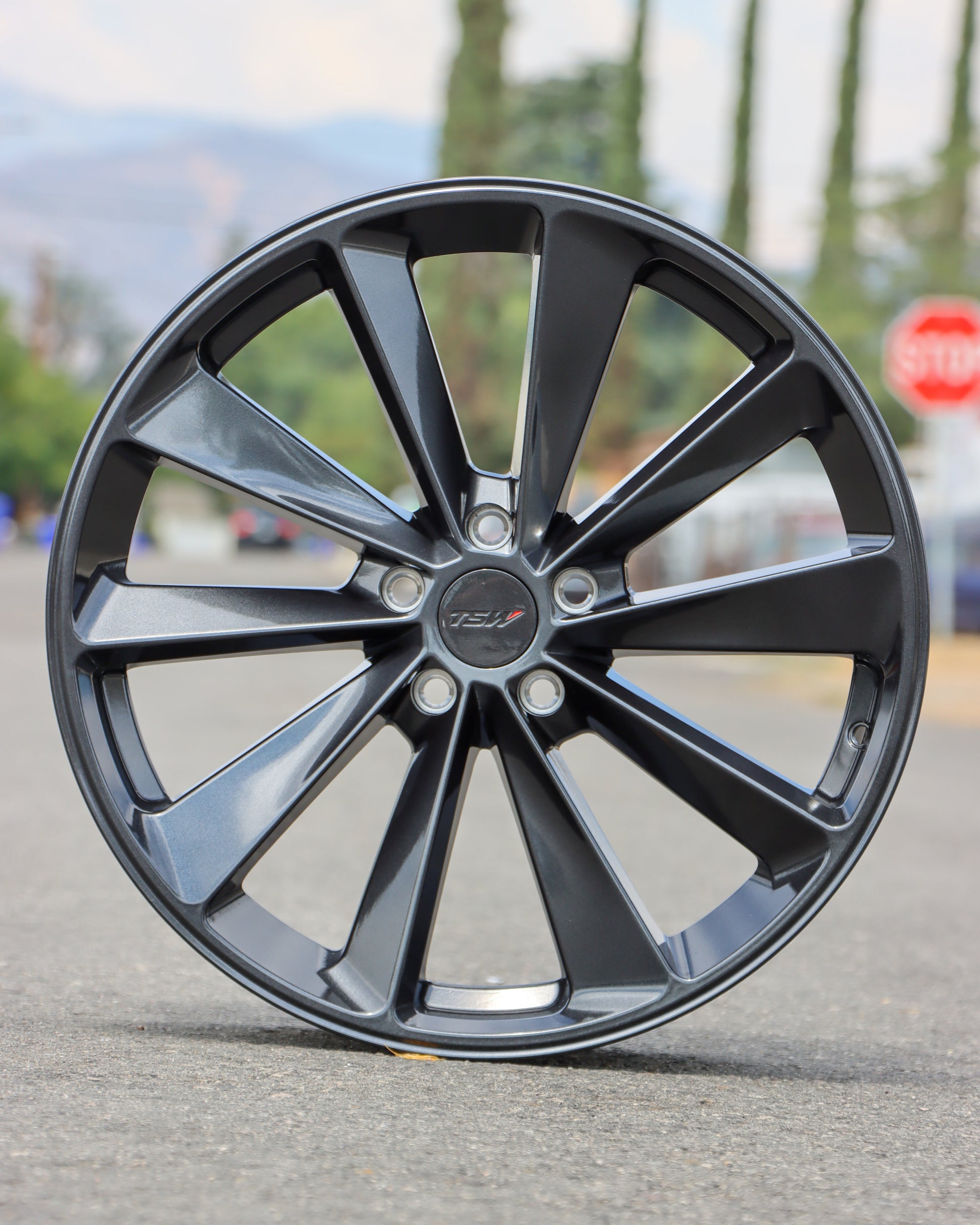 TSW Aileron Wheel in a gunmetal gray finish Sitting in the middle of the street with trees and a stop sign in the background.