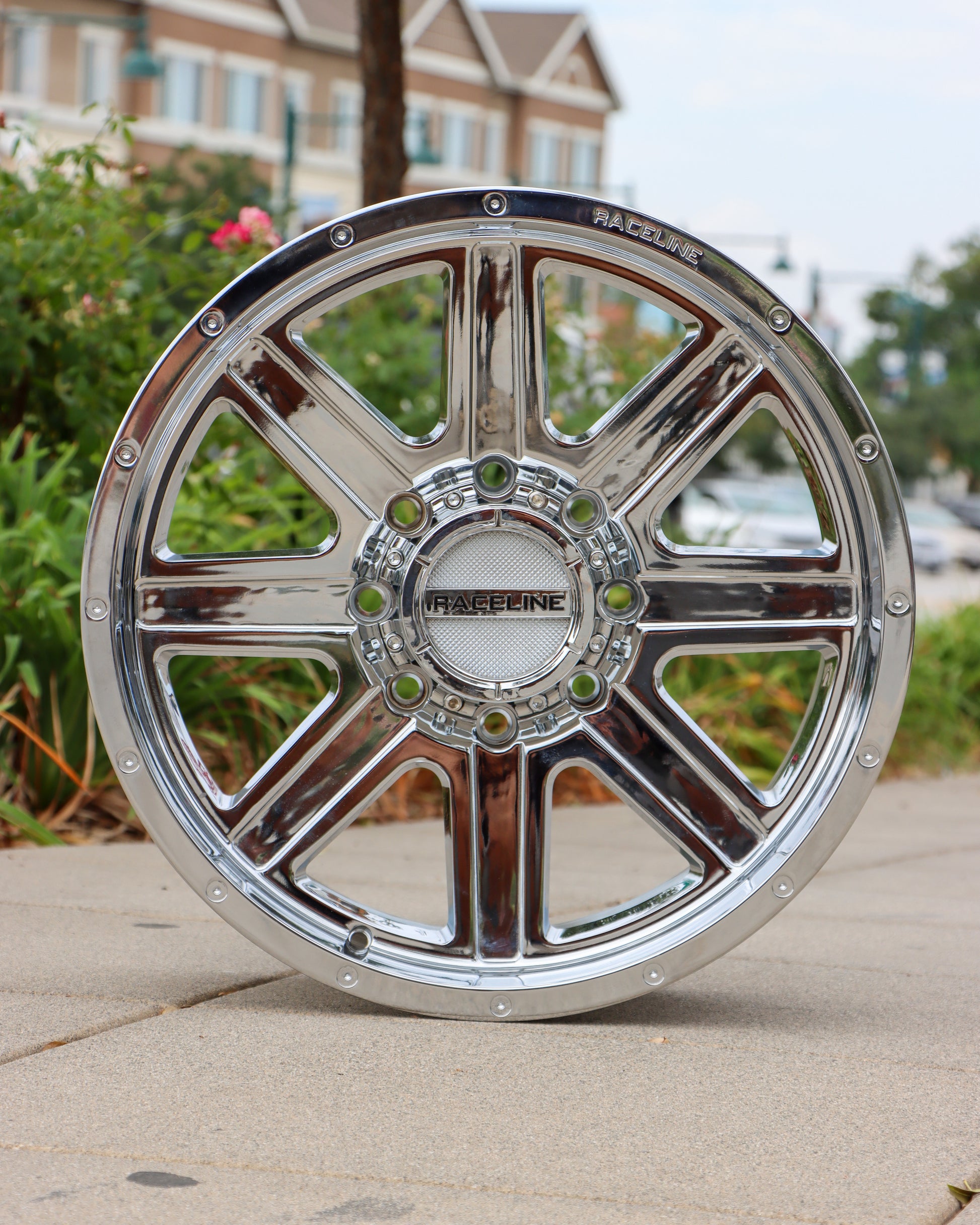 Raceline HOstage wheel in a chrome finish sitting on the sidewalk with bushes and an apartment building in the background.