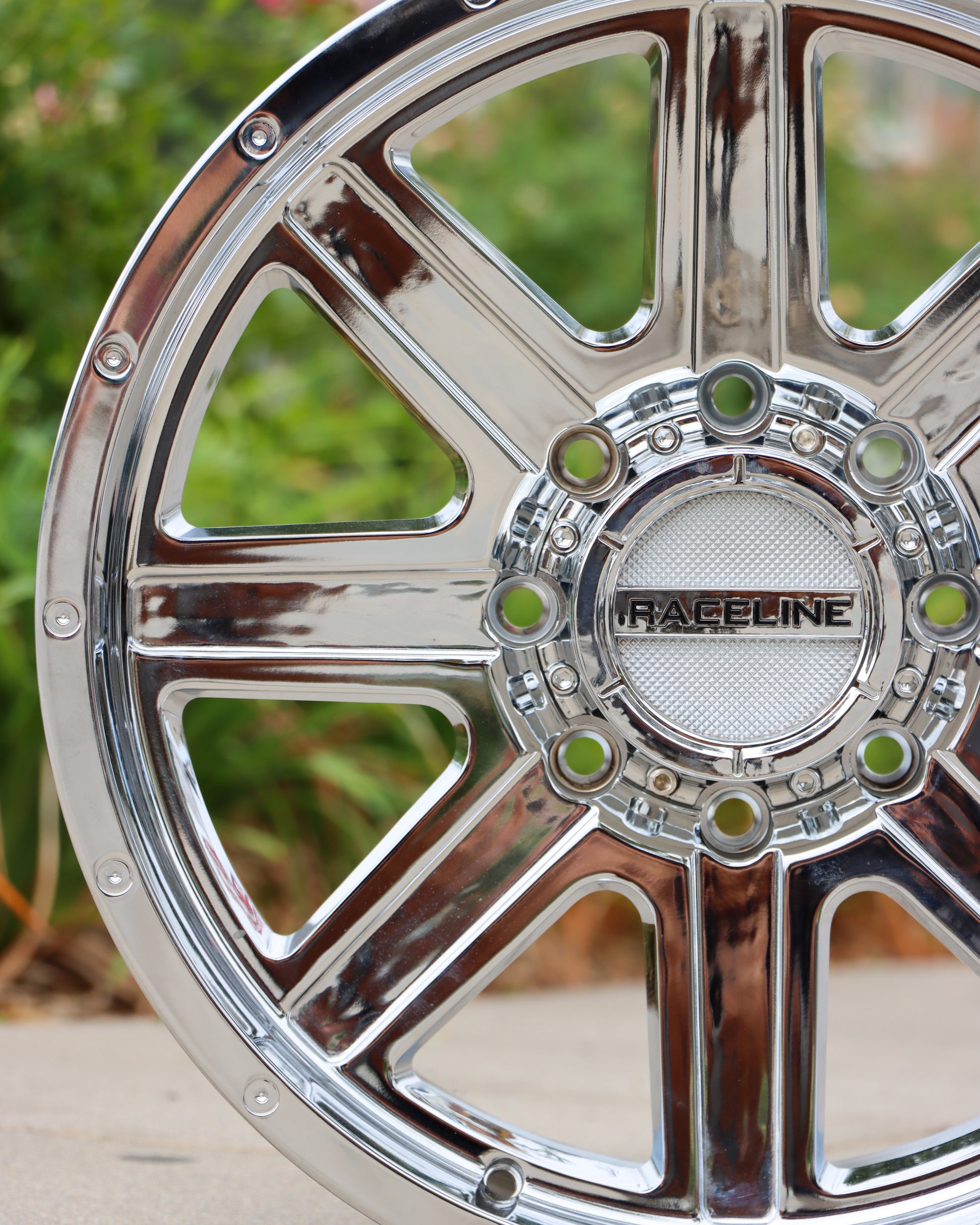 close-up view of the raceline hostage wheel in a chrome finish with bushes in the background.