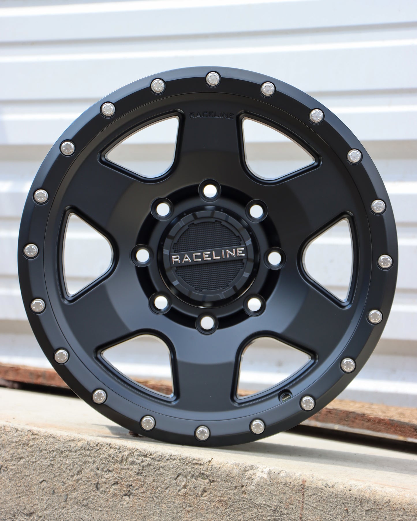 Raceline Boost wheel in a matte black finish sitting on some concrete with a metal sheet in the background.